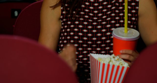 Person holding popcorn and drink while sitting in a movie theater. Ideal for content about entertainment, hobbies, relaxation, and leisure activities or promoting movie theaters and cinema experiences.