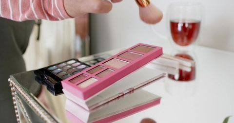 African american woman reaching for pink eyeshadow palette on mirrored vanity with tea cup