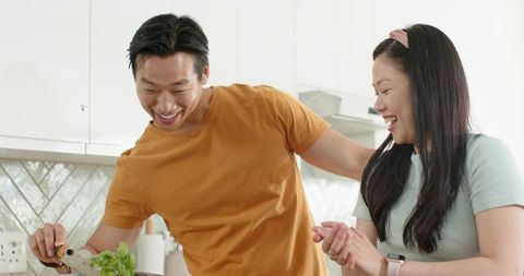 Asian Couple Cooking Together Pouring Oil into Pan Smiling in Modern Sunlit Kitchen