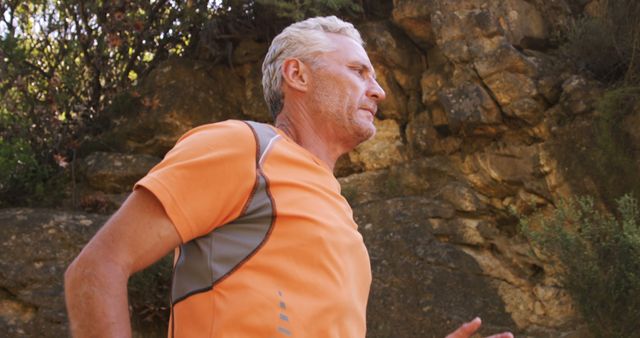 This image can be used to promote healthy living and fitness routines for seniors. Ideal for websites, magazines, or advertisements focusing on exercise, wellness, and outdoor activities. The vibrant orange shirt contrasts well against the rocky natural background, suggesting energy and vitality.