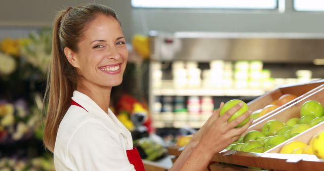 Female grocery store worker smiling while arranging green apples in the produce section. Ideal for use in retail, supermarket promotions, advertisements, healthy eating campaigns, and customer service training materials.