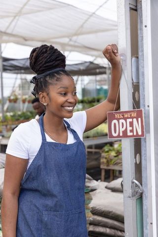 Smiling african american woman holding yes we are open sign at greenhouse plant nursery