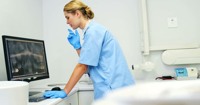 Female dentist wearing blue scrubs and gloves, attentively observing dental x-rays on computer screen. Modern dental clinic with medical equipment in background. Ideal for healthcare publications, dental care advertisements, or medical training materials.