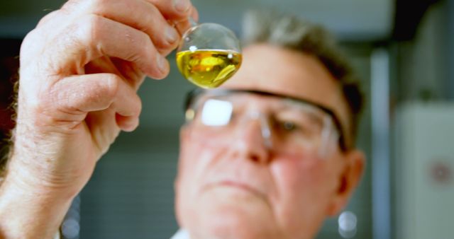 Scientist wearing safety goggles concentrating while holding up beaker containing liquid sample, ideal for depicting scientific research, laboratory work, chemistry experiments, or medical testing environments.