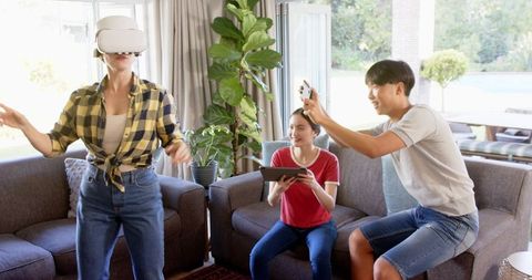 Asian woman using VR headset at home while friends capture reactions on tablet and phone