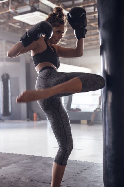 Caucasian dedicated young female boxer wearing black gloves kicking punching bag in health club. Practicing, unaltered, boxing, sport, training, strength and fitness concept.