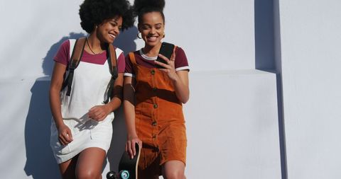Smiling twins relaxing with smartphone and skateboard on sunny day