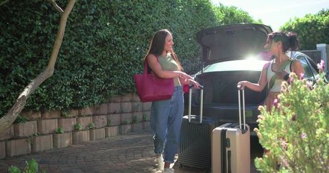 Two women loading suitcases into car trunk before road trip, smiling friends with bright tote