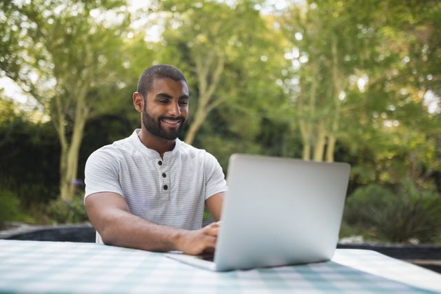 Man sitting at a table on a patio, using a laptop and smiling. Surrounded by trees and greenery, creating a relaxed and natural atmosphere. Ideal for illustrating concepts of remote work, freelancing, outdoor activities, and casual technology use.