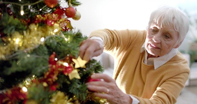 The image portrays an elderly woman decorating a Christmas tree with colorful ornaments and lights, embodying a festive holiday spirit. This can be used in holiday promotions, greeting cards, and senior living articles, as well as content related to Christmas traditions and family celebrations.