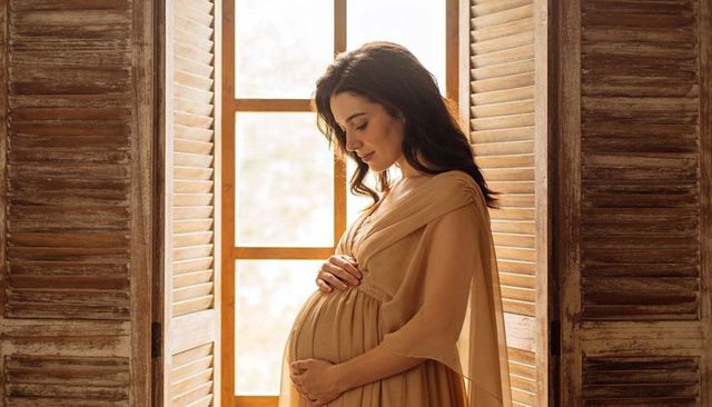 Expectant mother cradling belly in sunlit rustic room with wooden shutters