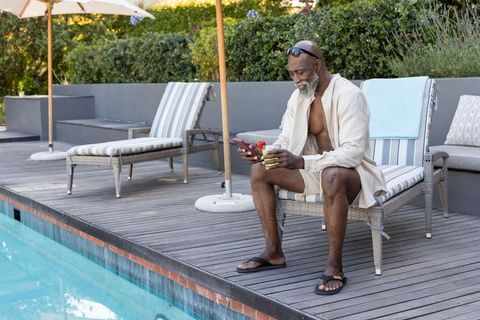 Mature african american man relaxing poolside on chaise lounge with smartphone and cocktail
