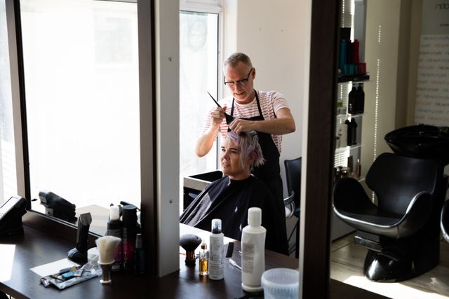 Middle aged Caucasian male hairdresser coloring a young Caucasian woman's hair in a salon, reflected in a mirror. Ideal for use in articles or advertisements related to beauty, hair care, professional hairstyling services, and salon environments.