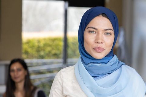 Confident woman wearing layered blue hijab standing in office lobby with colleague behind
