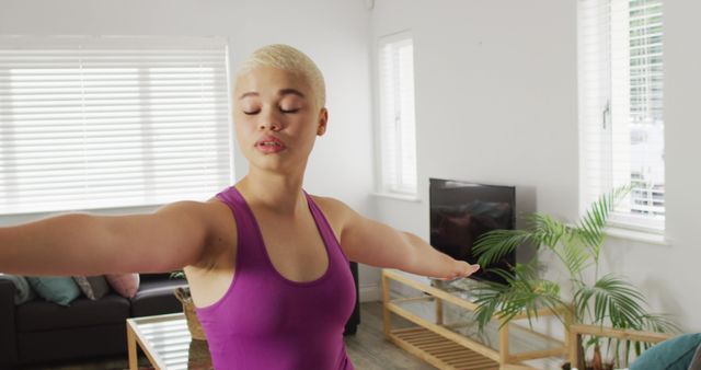 A young woman in a brightly lit living room practicing yoga. She is focused, with her eyes closed, showing balance and concentration. The indoor setting with modern decor creates a calm and peaceful atmosphere, ideal for promoting fitness and wellness. Perfect for use in websites and publications about home workouts, fitness tips, mindfulness practices, and healthy living.