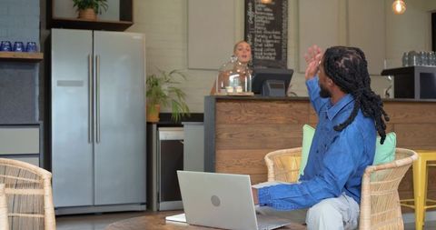 Remote worker waving to barista in cozy cafe workspace with laptop and wicker seating