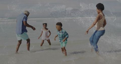 Family of four splashing in shallow sea on overcast beach, parents playing with kids