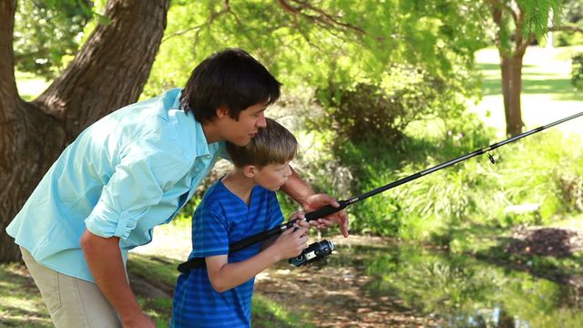 A father is teaching his young son how to fish in a picturesque countryside setting. Lush greenery envelops them, creating a serene backdrop for the bonding experience. Ideal for topics related to family activities, outdoor leisure, parenting, and learning moments. It can be used for promotions about summer vacations, family outing inspiration, or educational materials on outdoor skills.