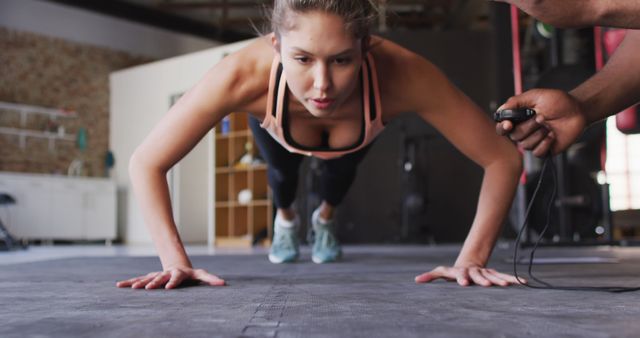 A focused woman in sportswear performing push-ups on a gym floor while her personal trainer times her performance. Ideal for use in fitness, training programs, workout tutorials, motivational content, and healthy lifestyle promotions.