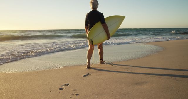 This image features a senior man standing on the beach while holding a surfboard, gazing at the ocean during sunset. Ideal for promoting active aging, retirement life, water sports, and coastal living. Useful for travel brochures, wellness campaigns, and beach resort advertisements.