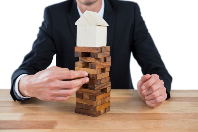 Businessman arranging wooden blocks with a house model on top, symbolizing strategic planning, risk management, and stability in real estate or investment. Ideal for use in articles, presentations, or advertisements related to business strategy, real estate investment, and decision-making processes.