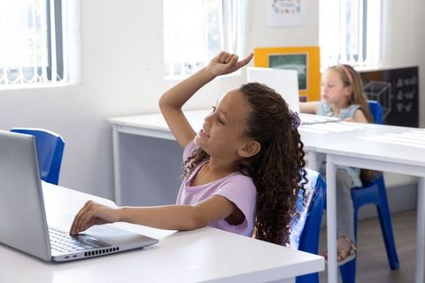 Girl raising hand while using laptop in classroom, engaged learning and digital education