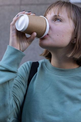 Young woman drinking takeaway coffee from paper cup in teal sweatshirt, closeup urban portrait