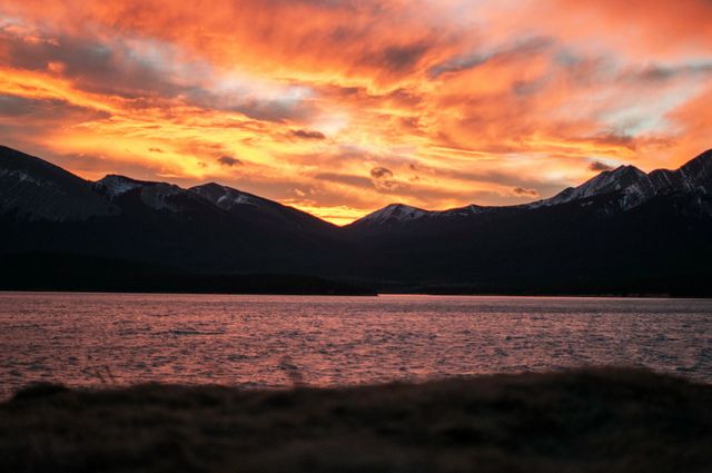 The vibrant colors of the sunrise illuminate the sky behind the silhouettes of the mountains, reflecting on the calm lake. Ideal for backgrounds, nature enthusiasts, travel brochures, or meditation content.