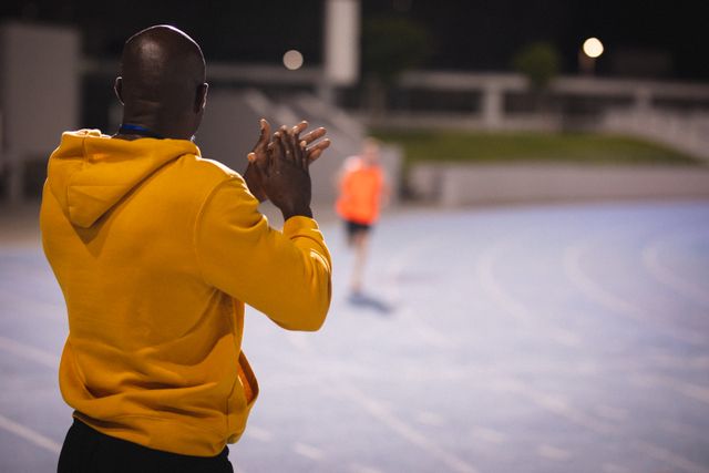Coach clapping hands while encouraging a runner during night training at a sports stadium. Ideal for use in articles about athletic training, motivation, coaching techniques, and sports events. Suitable for fitness blogs, sports magazines, and motivational content.