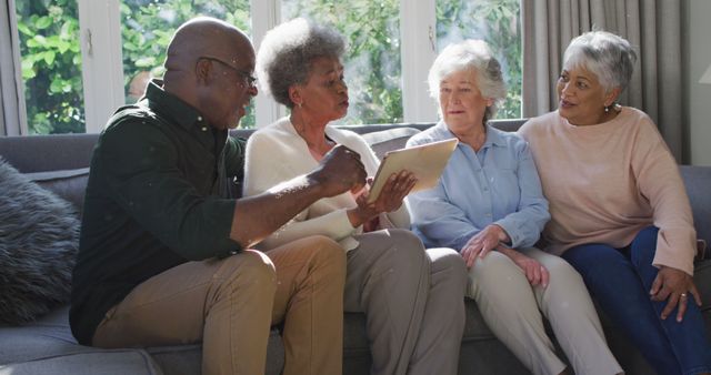 Group of seniors sitting on sofa in living room, engaging in conversation and looking at a tablet. Perfect for illustrating the use of technology among elderly, friendship in older age, and social gatherings. Useful for advertisements or articles related to senior living, technology for seniors, and social health.