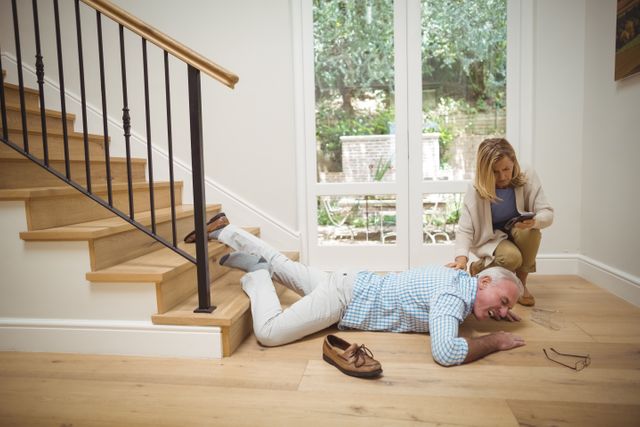Woman checking her mobile phone while a senior man has fallen downstairs at home. The scene depicts a potential emergency situation involving an elderly person. Useful for illustrating topics related to home safety, elderly care, accidents, and emergency response.