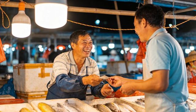 Smiling fish vendor accepting cash at night market stall with fresh seafood and coolers