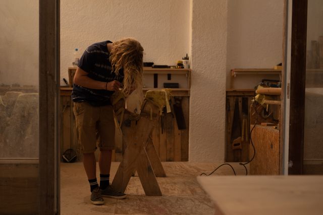 Caucasian male surfboard maker with long blonde hair working in his studio, standing, bent over a surfborad on a workbench.