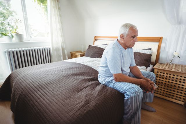 Senior man sitting on bed in bedroom, wearing pajamas, looking thoughtful. Ideal for topics related to aging, retirement, solitude, morning routines, and peaceful home environments.
