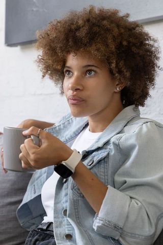 Sitting on sofa holding mug and reflecting, young woman in denim jacket, cozy portrait