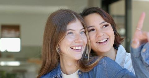 Smiling diverse female friends sharing joyful moment at home near window, casual denim lifestyle