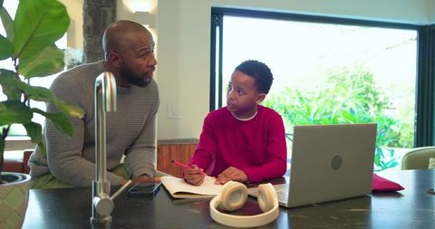 African american father guiding son doing homework at kitchen island with laptop and headphones