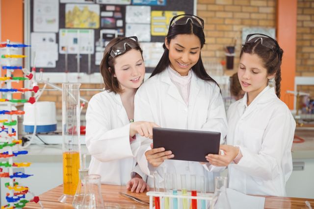 Three school girls wearing lab coats and safety goggles are using a digital tablet in a science laboratory. They are surrounded by test tubes and scientific equipment, indicating they are engaged in a chemistry experiment. This image is ideal for educational content, promoting STEM education, and illustrating teamwork and learning in a classroom setting.