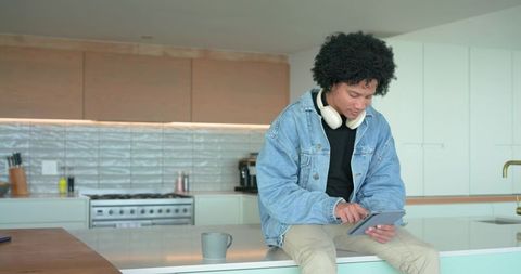 African American man sitting on kitchen island using tablet with headphones in modern home