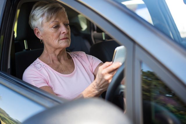 Senior woman sitting in driver's seat of car, using mobile phone. Ideal for illustrating topics related to road safety, distracted driving, elderly drivers, and the use of technology by seniors. Can be used in articles, advertisements, and educational materials promoting safe driving practices and awareness.