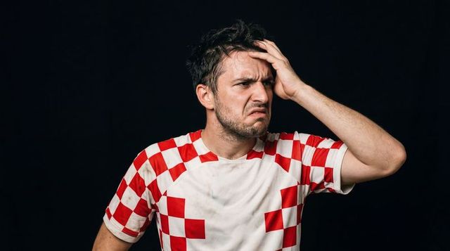 Frustrated soccer fan clutching forehead in stained red-white checkered jersey, closeup
