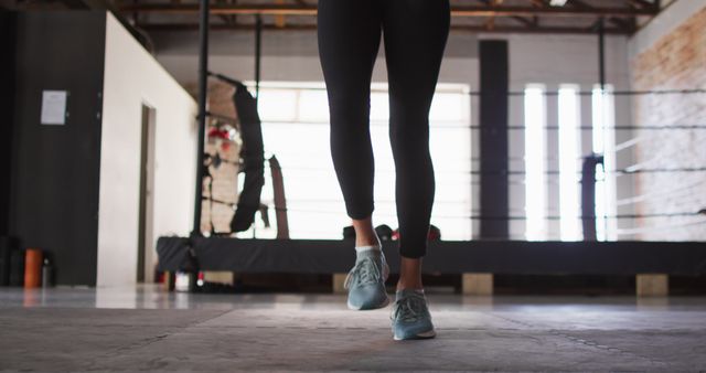 Legs of an athlete jumping rope on a gym floor. Person wearing athletic leggings and sneakers near a boxing ring. Useful for fitness blogs, workout guides, sports equipment advertisements, and exercise program promotions.