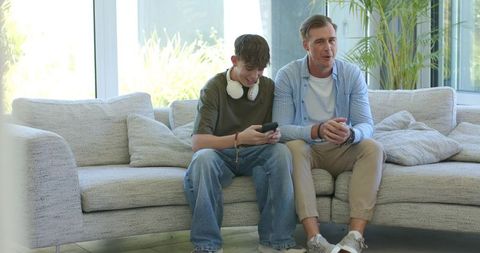 Father and teen son sharing moment on gray sectional sofa, teen scrolling smartphone