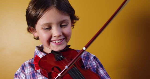 Young boy smiling while playing violin against solid yellow background, dressed in casual plaid shirt. Suitable for educational content, music school promotions, childhood development projects, and joyful moments of learning and play.
