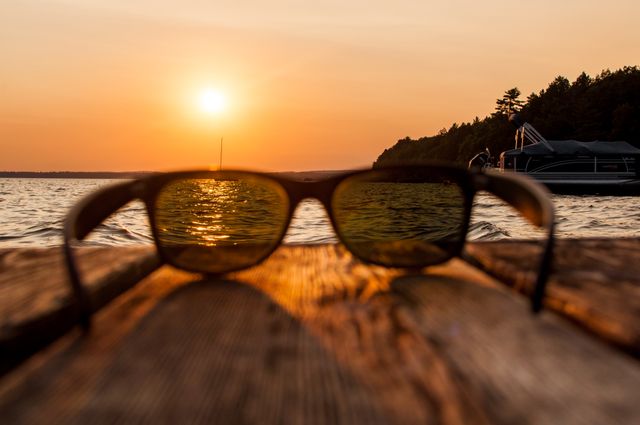 Close-up of sunglasses on a weathered wooden table with a serene lake and setting sun in the background. Ideal for use in travel blogs, vacation advertisements, and leisure-themed content.