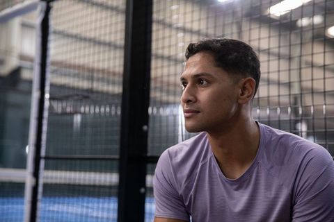 Asian athlete resting on bench beside wire mesh court, looking focused and thoughtful