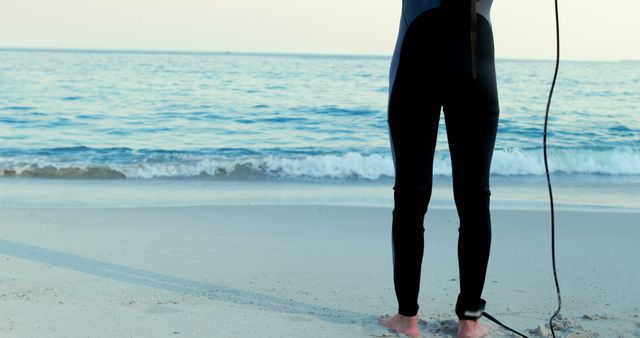 Surfer in black wetsuit standing on sandy beach near ocean with surfboard leash wrapped around their ankle. Ideal for use in advertisements, articles, or websites promoting surfing, beach vacations, or water sports.