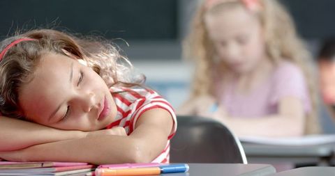 Girl wearing striped shirt sleeps on desk as classmate writes in background. Colorful pens beside her suggest classroom activity. Useful for topics on children's education, fatigue in learning environments, classroom management, and student behavior.