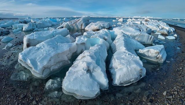 Blue icebergs melting on pebble shoreline, calm arctic sea, glacial landscape