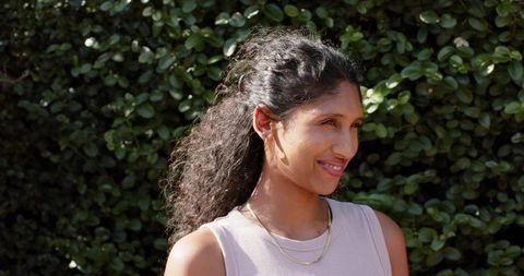Sunlit smiling woman standing by green hedge wearing sleeveless top and gold necklaces